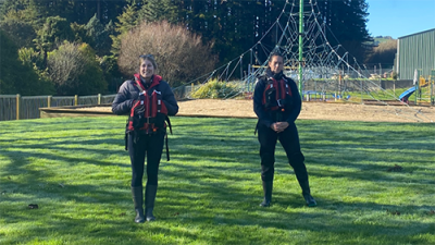 Image of two ladies standing on a field in wetsuits and lifejackets